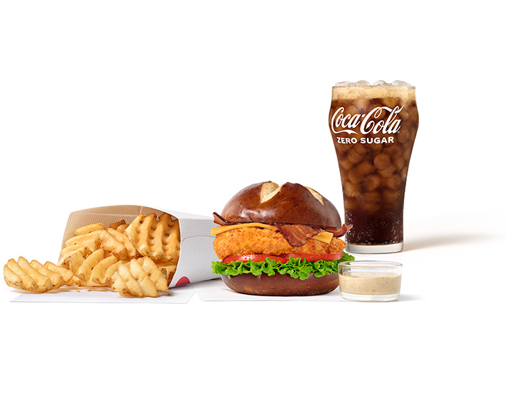 Pretzel Cheddar Club Sandwich with Original filet on white napkins next to a small glass bowl of Creamy Dijon Mustard Sauce, Waffle Fries, and a glass of Coca-Cola®