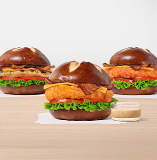 Three Pretzel Cheddar Club Sandwiches, one with the Original filet, one with the Grilled filet and one with Spicy filet all on white napkins next to a small glass bowl of Creamy Dijon Mustard Sauce