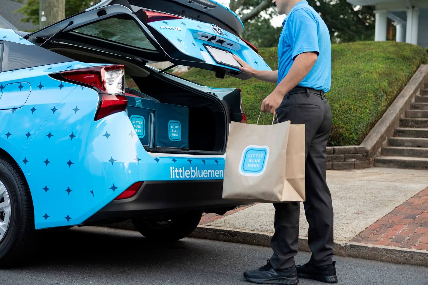 Little Blue Menu Team Member loads a bag full of food into the trunk of a blue branded Little Blue Menu delivery vehicle.  