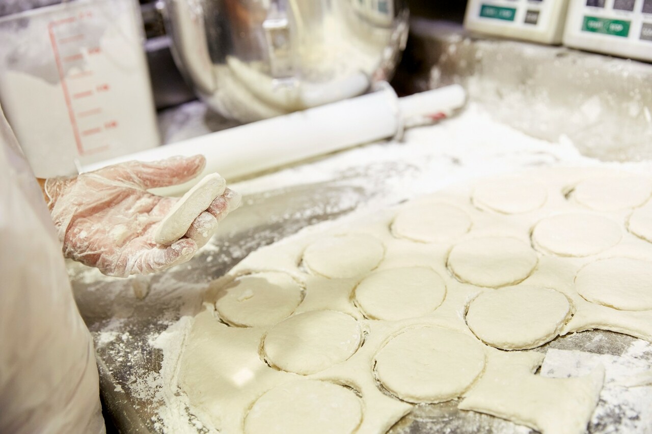 Chick-fil-A employee in plastic gloves uses a biscuit cutter on a sheet of dough to shape biscuits. 