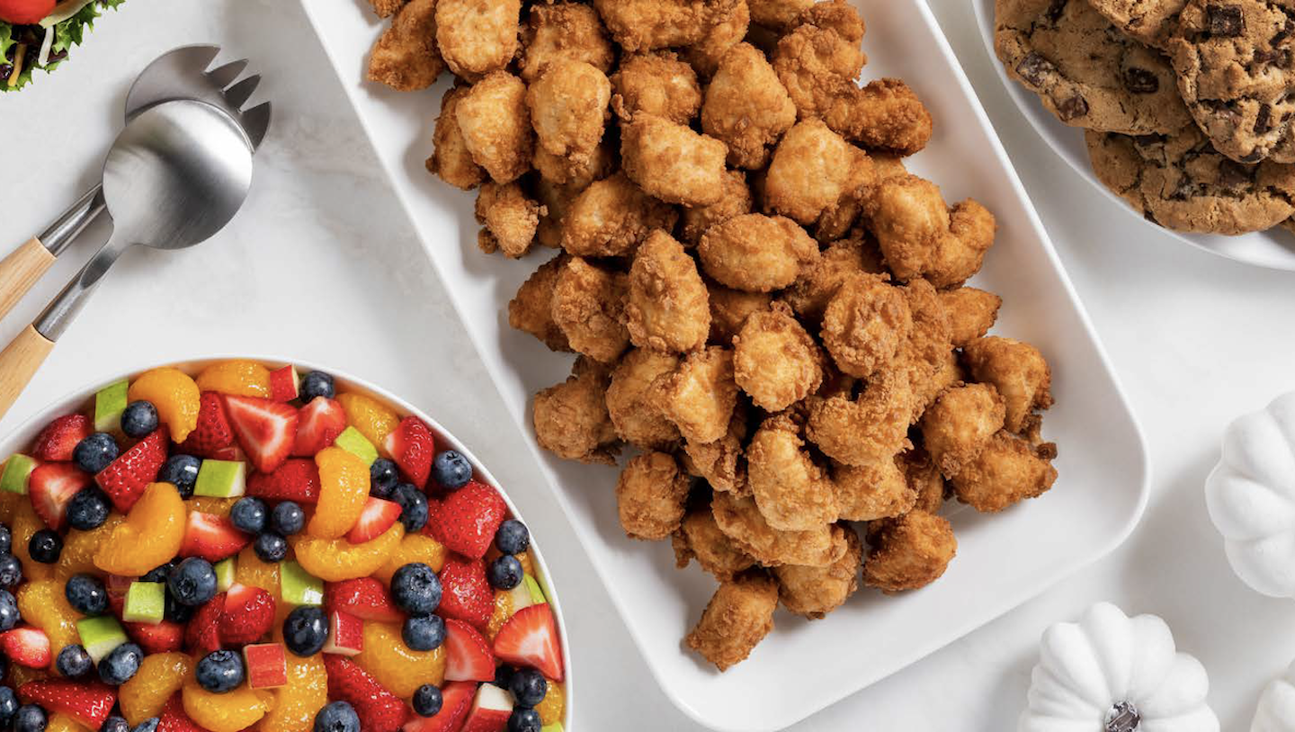 Items from Chick-fil-A’s catering menu, including a Fruit Tray, Chick-fil-A Nuggets Tray, Chocolate Chunk Cookie Tray and Side Salad are arranged on white dishes on a table. 