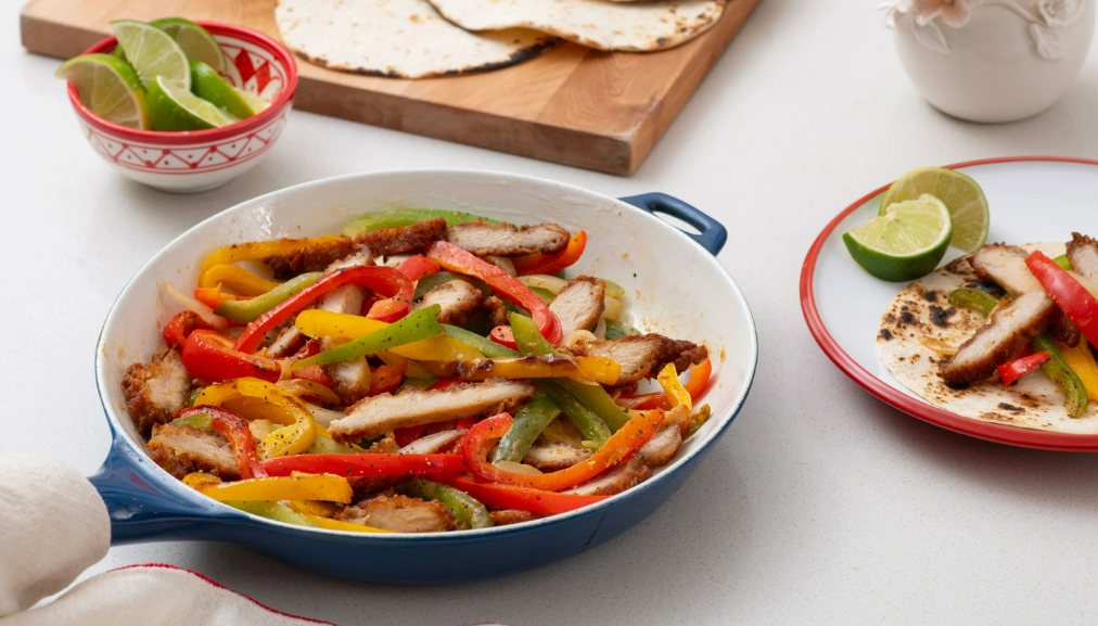 Skillet with Chick-fil-A Breaded Chicken Fajitas on a counter with serving board and plate in the background.