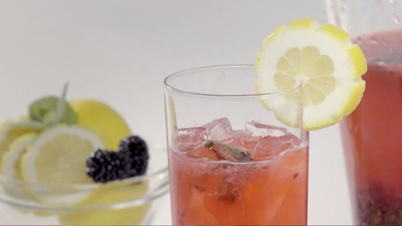 A glass of sparkling blackberry mint lemonade garnished with a lemon slice. A pitcher of lemonade and a bowl of fruit sit in the background.