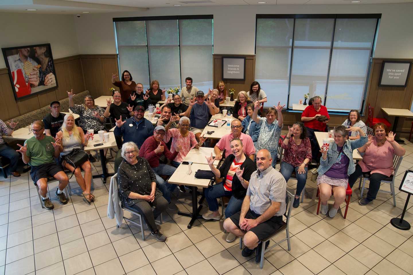 Participants of a Deaf Chat Night, bringing together both Deaf and hearing members of the community who communicate in American Sign Language over a meal, are seated in a Chick-fil-A restaurant. 