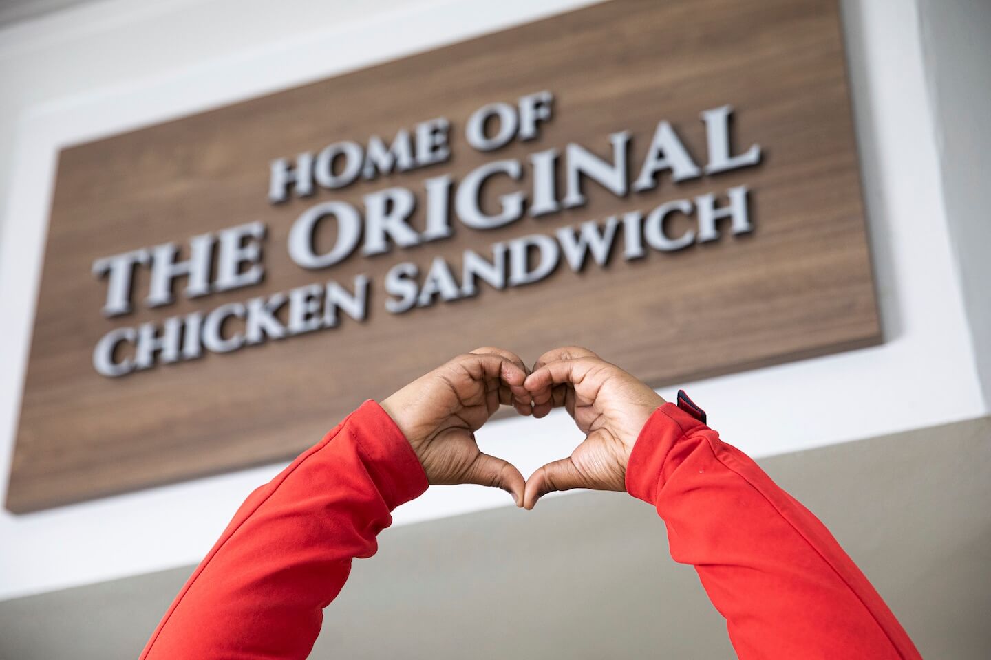 A Team Member holding a heart up with their hands in front of a sign reading "Home of the Original Chicken Sandwich.