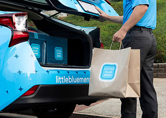 A blue Little Blue Menu delivery car and brown bag with Little Blue Menu logo.