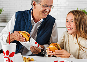 A father and daughter smiling while enjoying two Chick-fil-A Original Chicken Sandwiches.