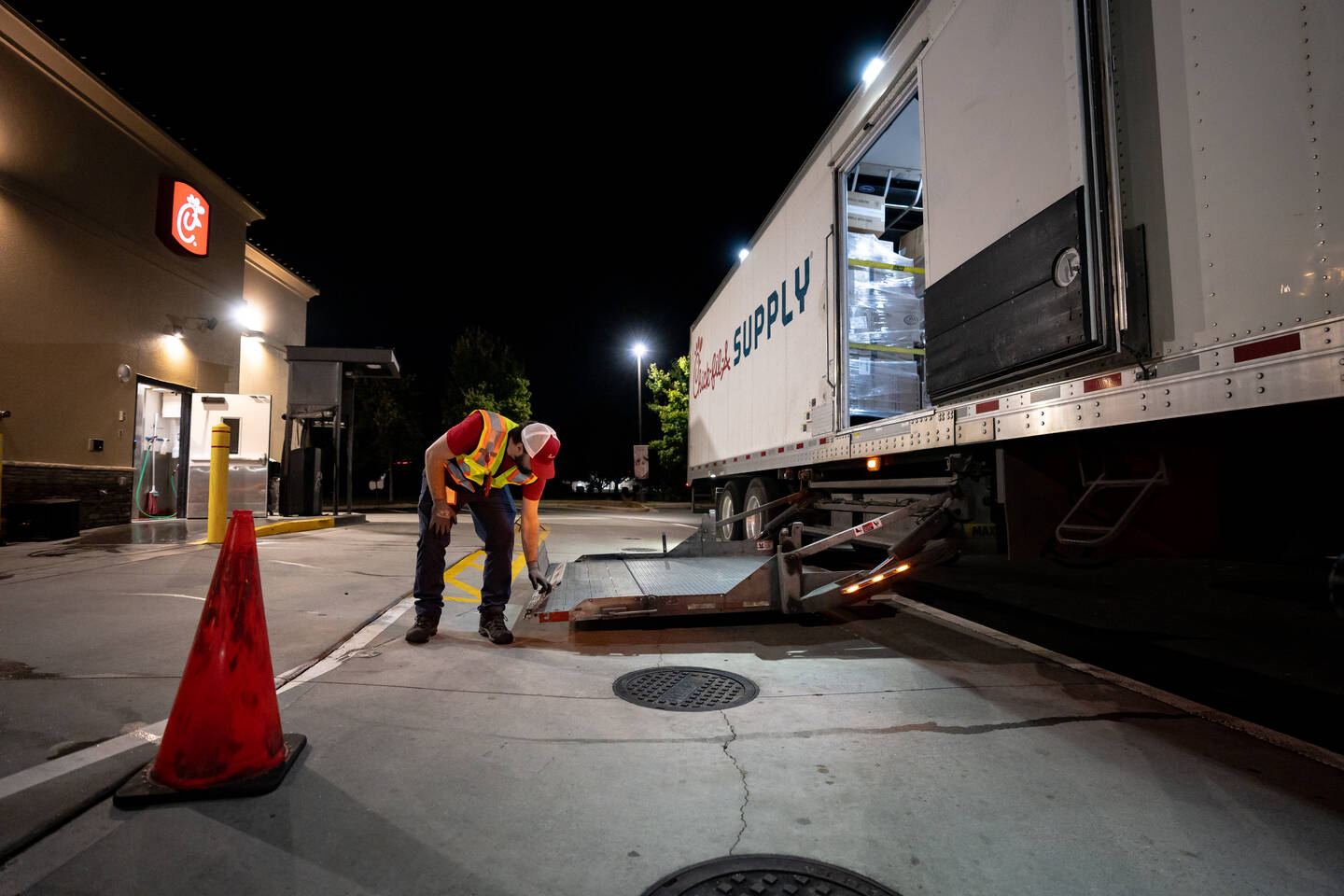 A man operating the ramp of a Chick-fil-A Supply Truck in a dark driveway next to a restaurant.