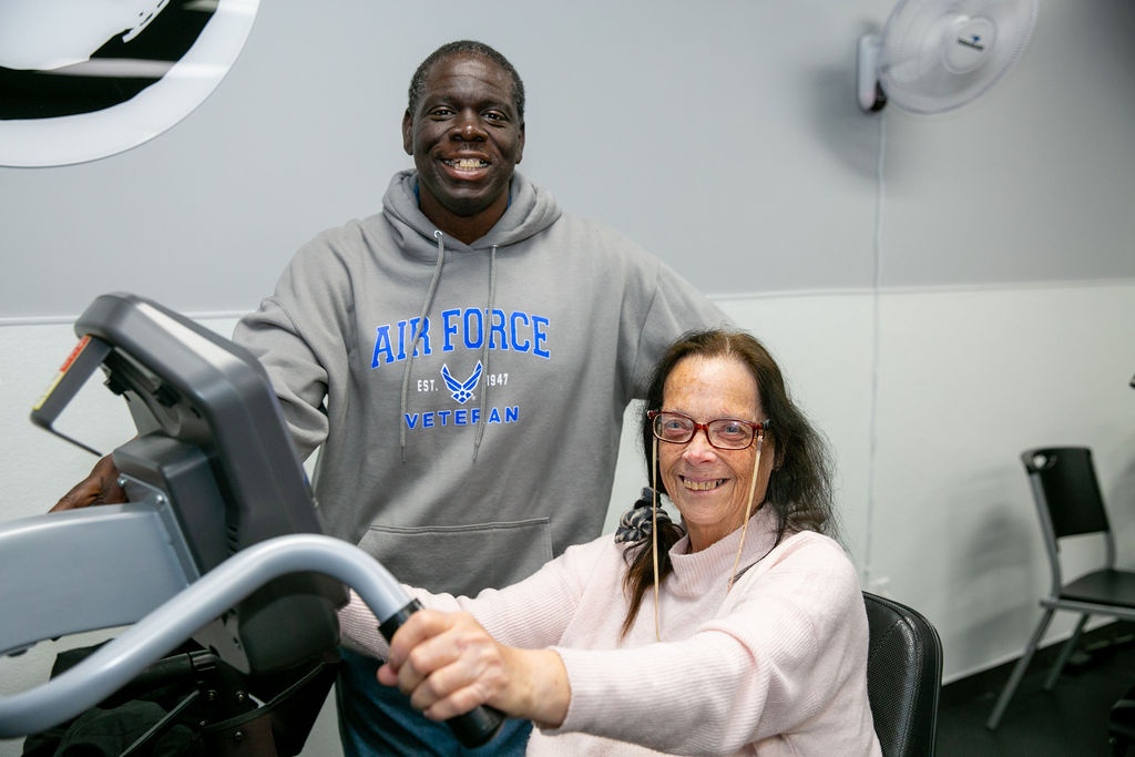 Woman sitting on a workout machine with man standing next to her smiling. 