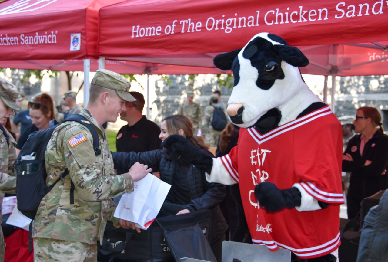 A member of the U.S. military in camouflage fatigues receiving a bag of food from a Chick-fil-A Cow mascot.