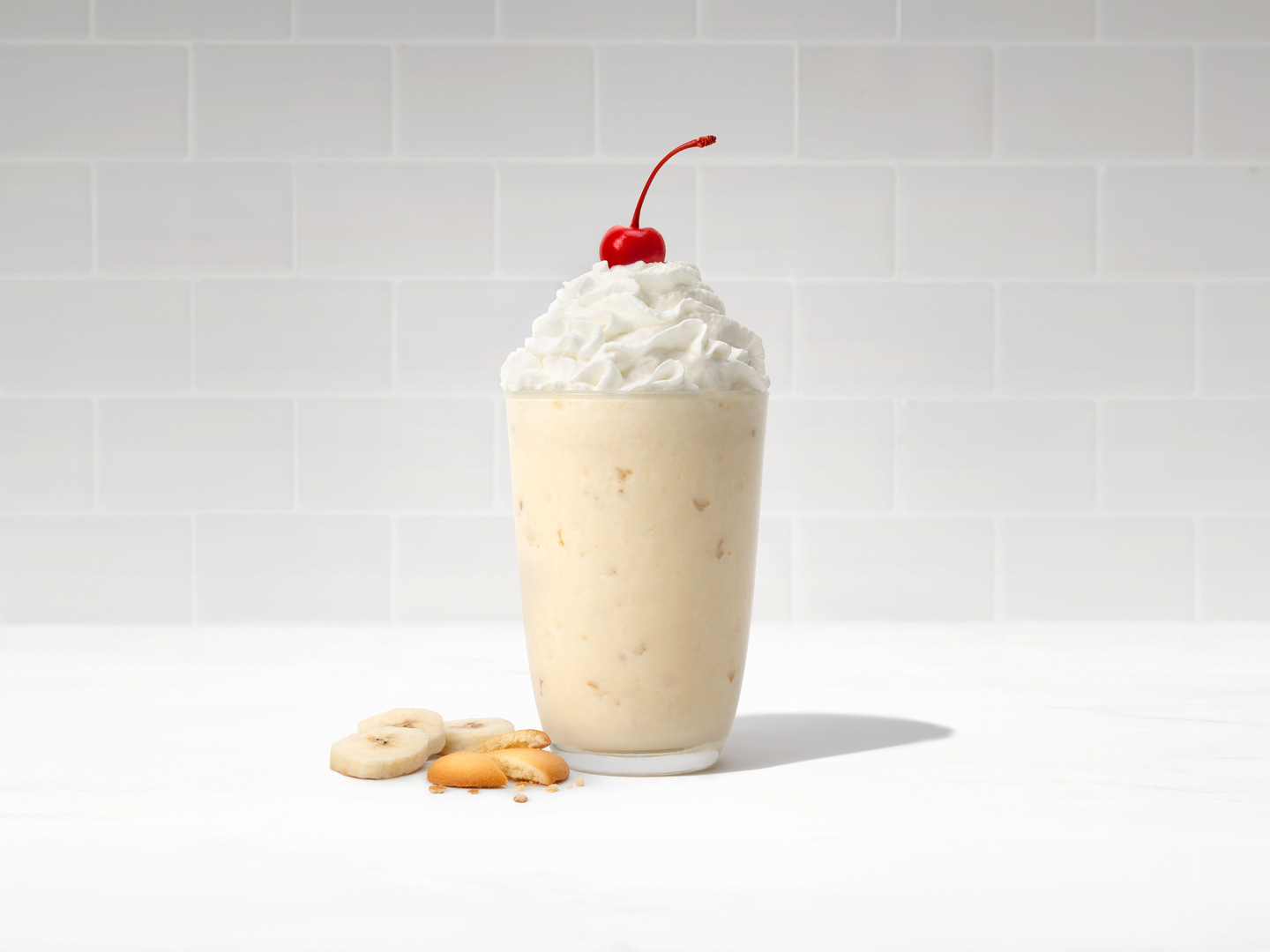 The new Chick-fil-A Banana Pudding Milkshake topped with whipped cream and a cherry in a glass on a white countertop next to three slices of banana and a vanilla wafer.