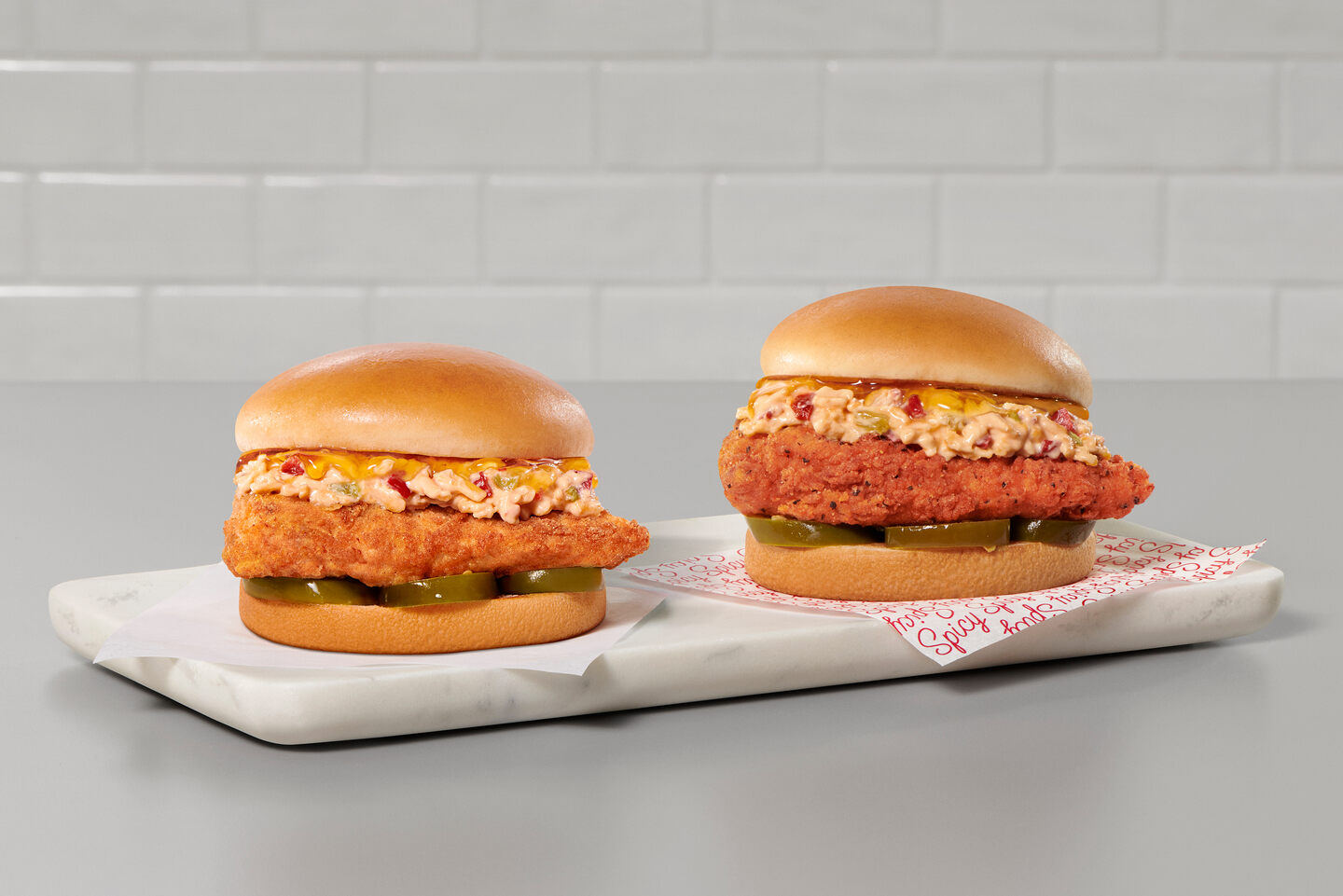 Two Chick-fil-A Honey Pepper Pimento Sandwiches on a granite serving board on top of a gray countertop, one with an Original fried filet and the other with a Spicy filet.