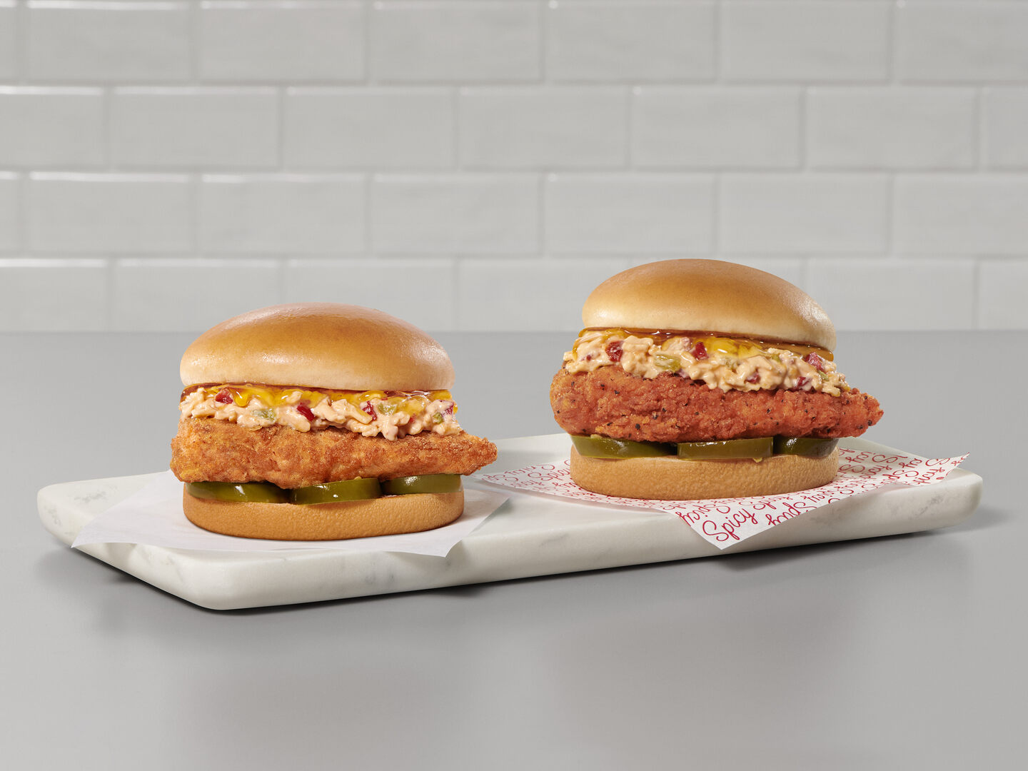 Two Chick-fil-A Honey Pepper Pimento Sandwiches on a granite serving board on top of a gray countertop, one with an Original fried filet and the other with a Spicy filet.