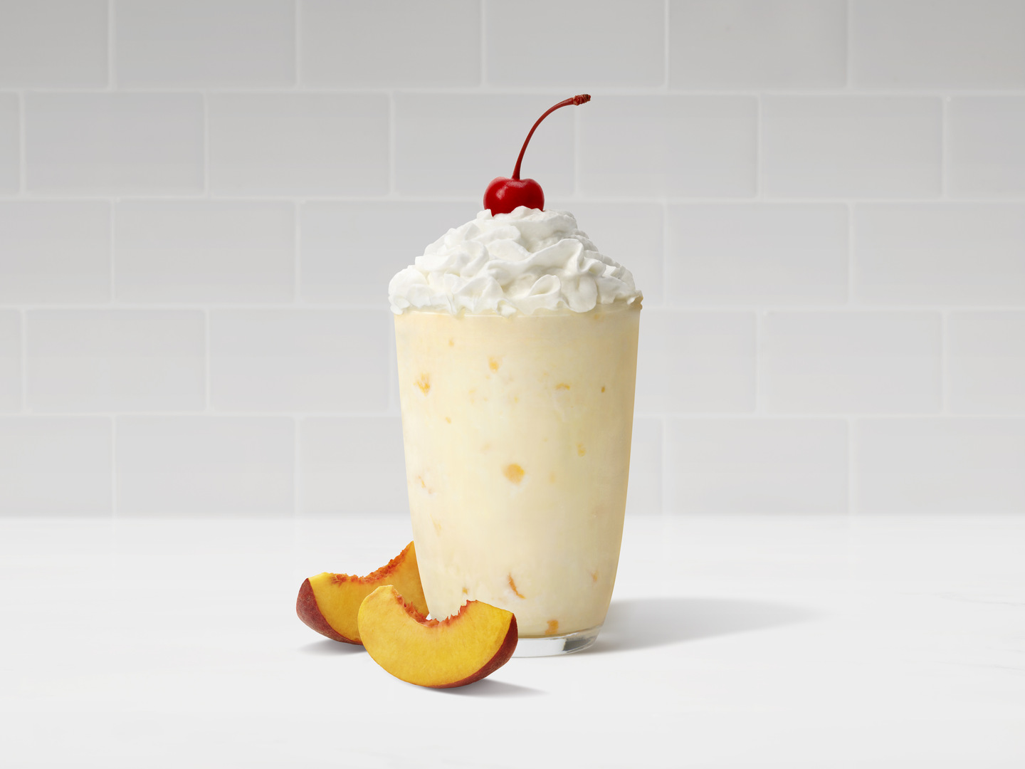 The iconic Chick-fil-A Peach Milkshake next to two slices of peaches on a white countertop.