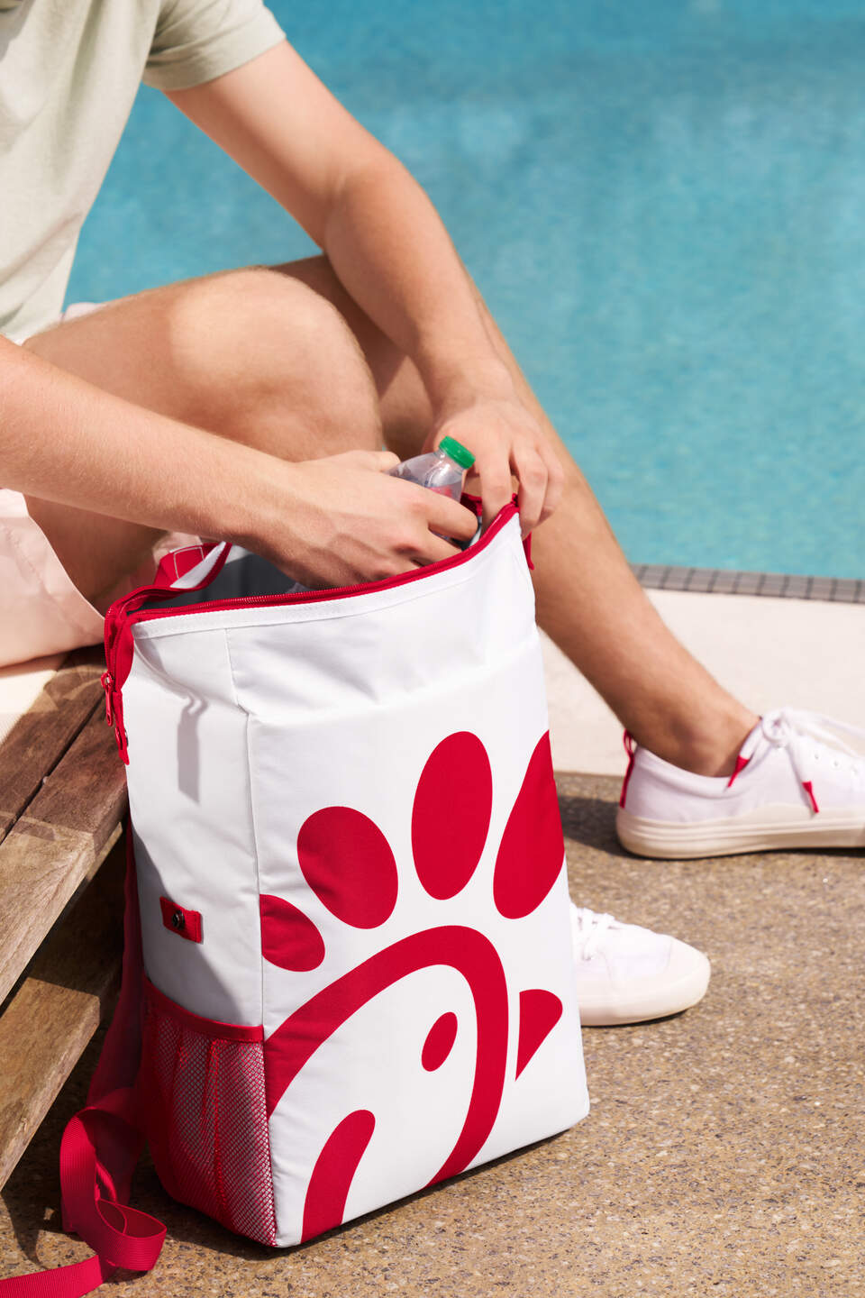 A male model putting a water bottle into the new Chick-fil-A Cooler Bag from the 2024 Summer Merchandise Launch while sitting next to a pool.