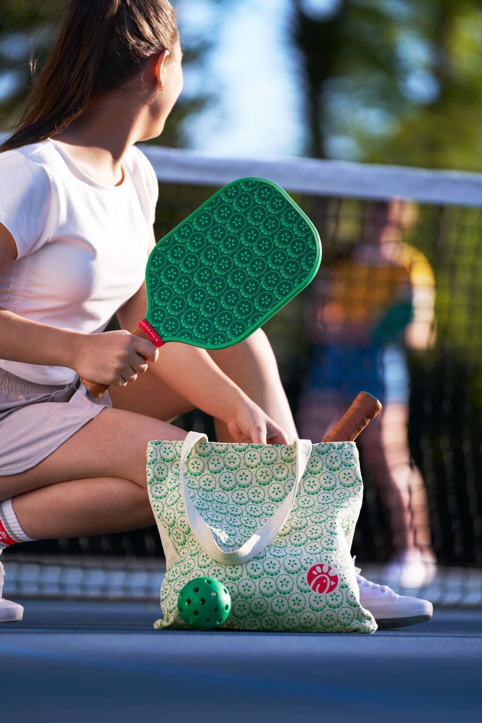 A woman kneeling next to a net on a pickleball court while holding the Chick-fil-A Pickleball Set from the 2024 Summer Merchandise Launch.