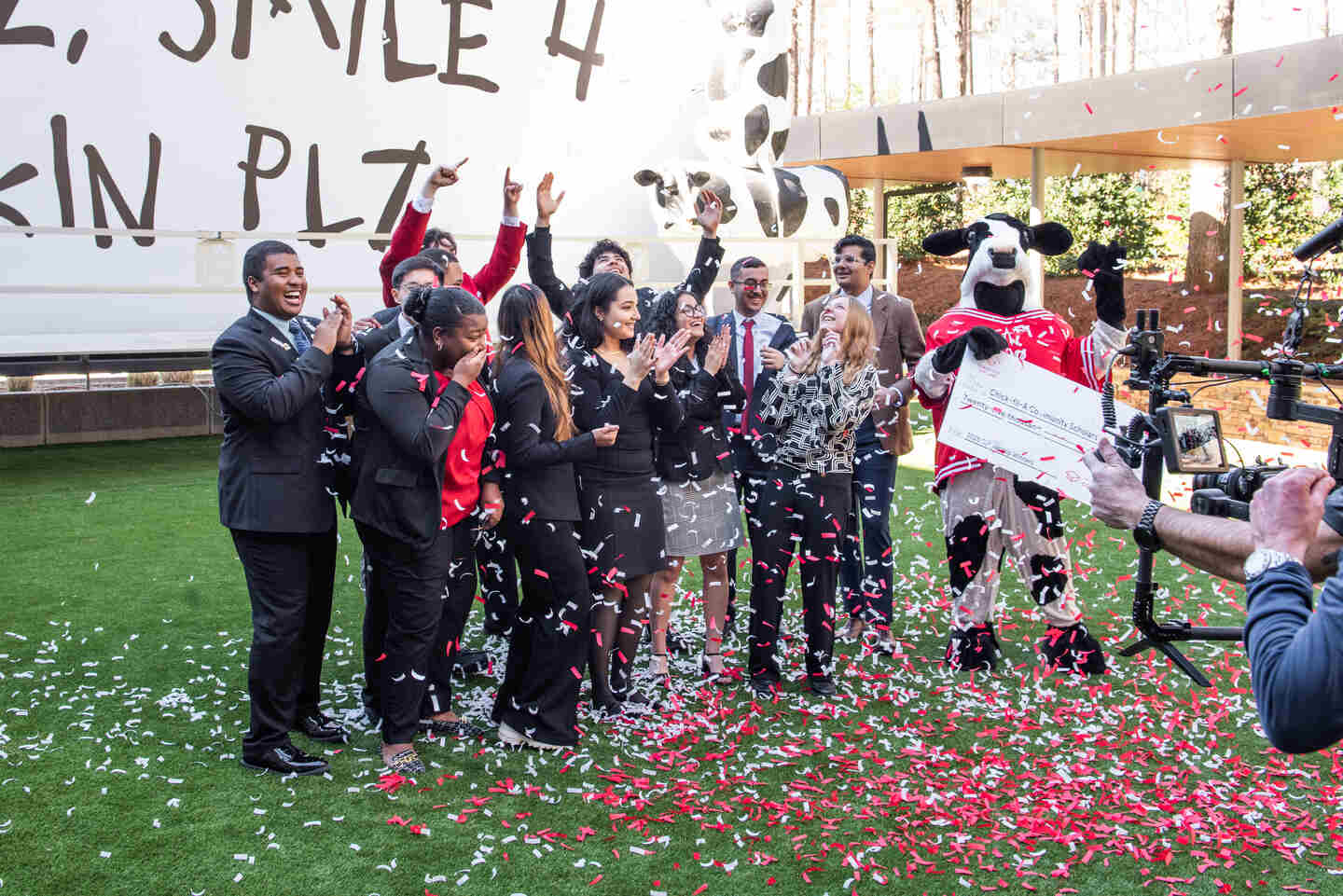 Group of Chick-fil-A Team Members celebrate receiving a Remarkable Futures Scholarship with red and white confetti, a camera crew, and the Chick-fil-A Cow mascot holding a large check.