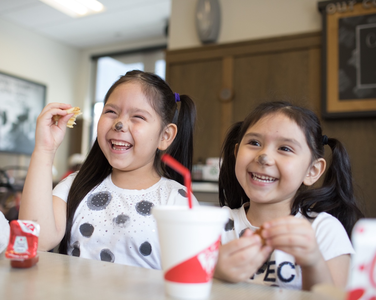 Two young girls with painted cow noses laugh while enjoying a meal at Chick-fil-A.