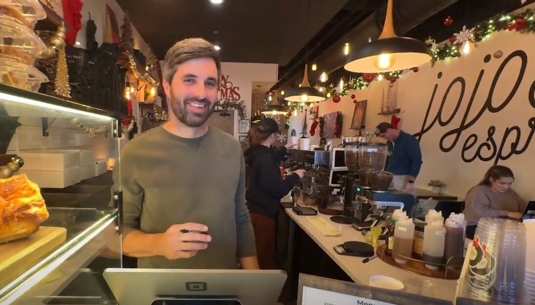 A smiling barista stands behind the counter at Jojo Espresso, a decorated café with warm lighting, pastries, and staff in the background.