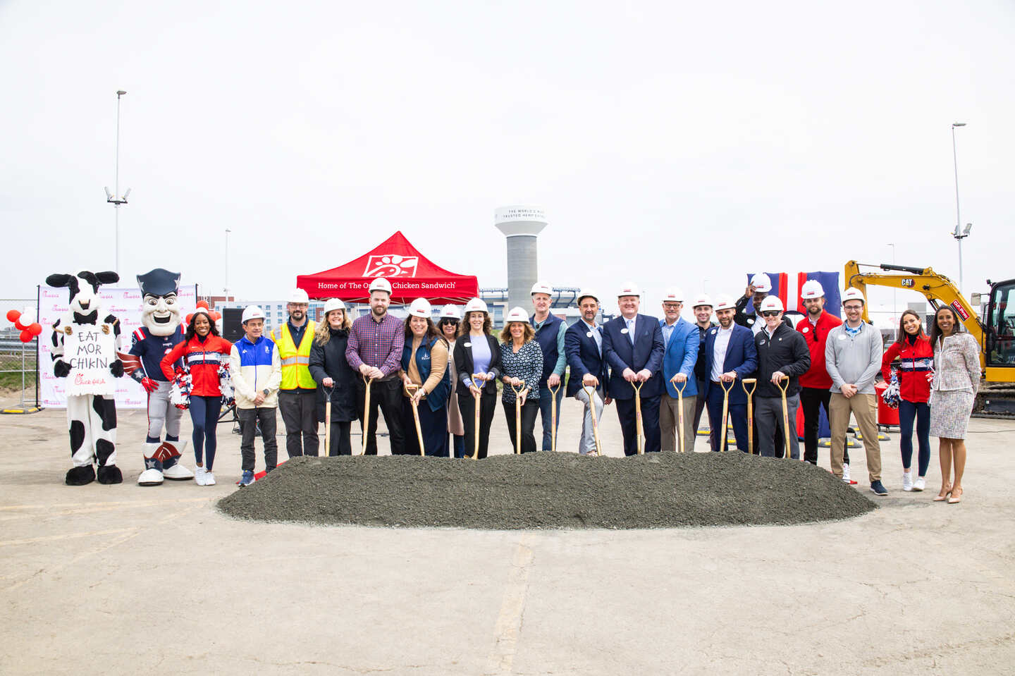 Chick-fil-A leaders, team members, and local partners pose with shovels at a groundbreaking ceremony, joined by the Chick-fil-A Cow and local mascots, in front of a red tent and construction equipment. 