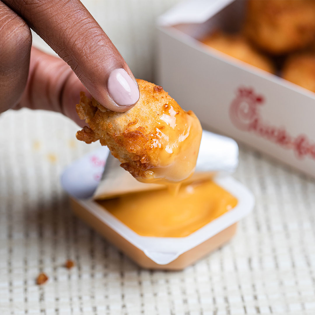 Person dipping a nugget into Chick-fil-A® Sauce with a package of Chick-fil-A® Nuggets in the background