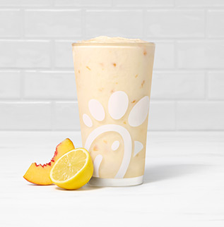 A glass of Peach Frosted Lemonade with a peach slice and a lemon half on a white countertop with a white tiled background