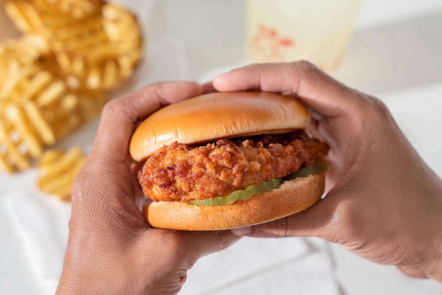 Person holding Chick-fil-A® Chicken Sandwich with Waffle Potato Fries® and Chick-fil-A® Lemonade in the background