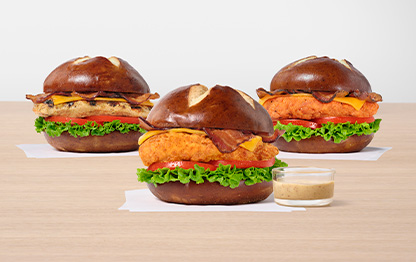 Three Pretzel Cheddar Club Sandwiches, one with the Original filet, one with the Grilled filet and one with Spicy filet all on white napkins next to a small glass bowl of Creamy Dijon Mustard Sauce