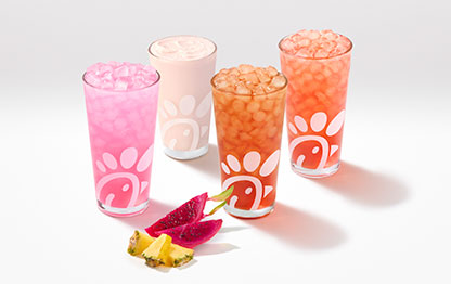 A glass of a Pineapple Dragonfruit Lemonade, Frosted Lemonade, Iced Tea and Sunjoy® with pieces of pineapple and dragonfruit next to them on a gray background