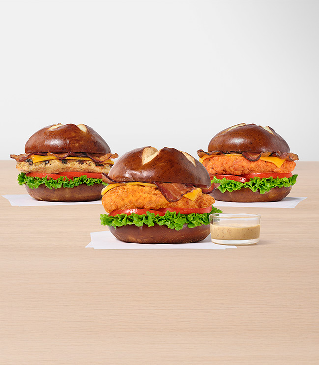 Three Pretzel Cheddar Club Sandwiches, one with the Original filet, one with the Grilled filet and one with Spicy filet all on white napkins next to a small glass bowl of Creamy Dijon Mustard Sauce