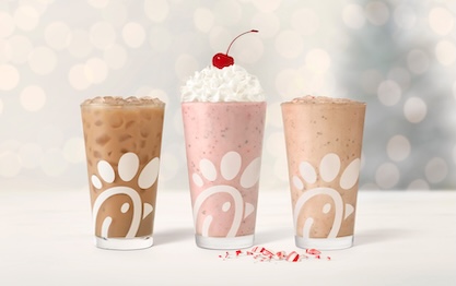 Glasses of Peppermint Chip Milkshake, Peppermint Iced Coffee, and Peppermint Chip lined up in a row, sitting next to pieces of peppermint, with twinkling holiday lights in the background