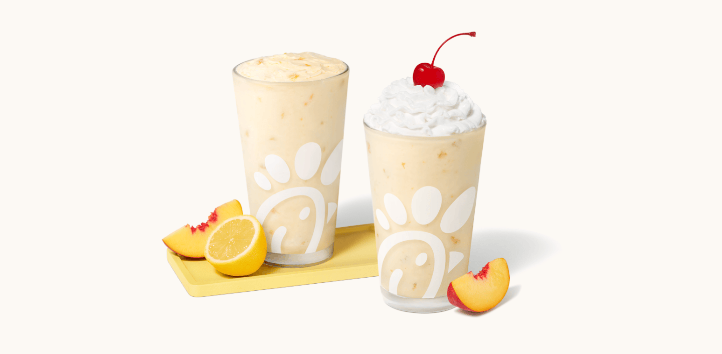 Peach Frosted Lemonade in a glass with a peach slice and a lemon half next to it, sitting next to a Peach Milkshake in a glass with a peach slice next to it