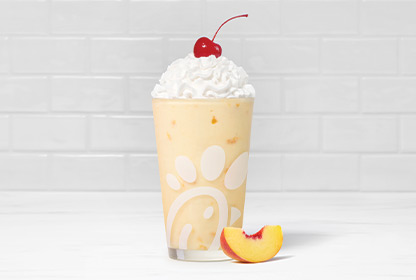 A glass of a Peach Milkshake with a peach slice next to it on a white countertop on a white tiled background