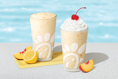 Peach Frosted Lemonade in a glass with a peach slice and a lemon half next to it, sitting next to a Peach Milkshake in a glass with a peach slice next to it with a pool in the background
