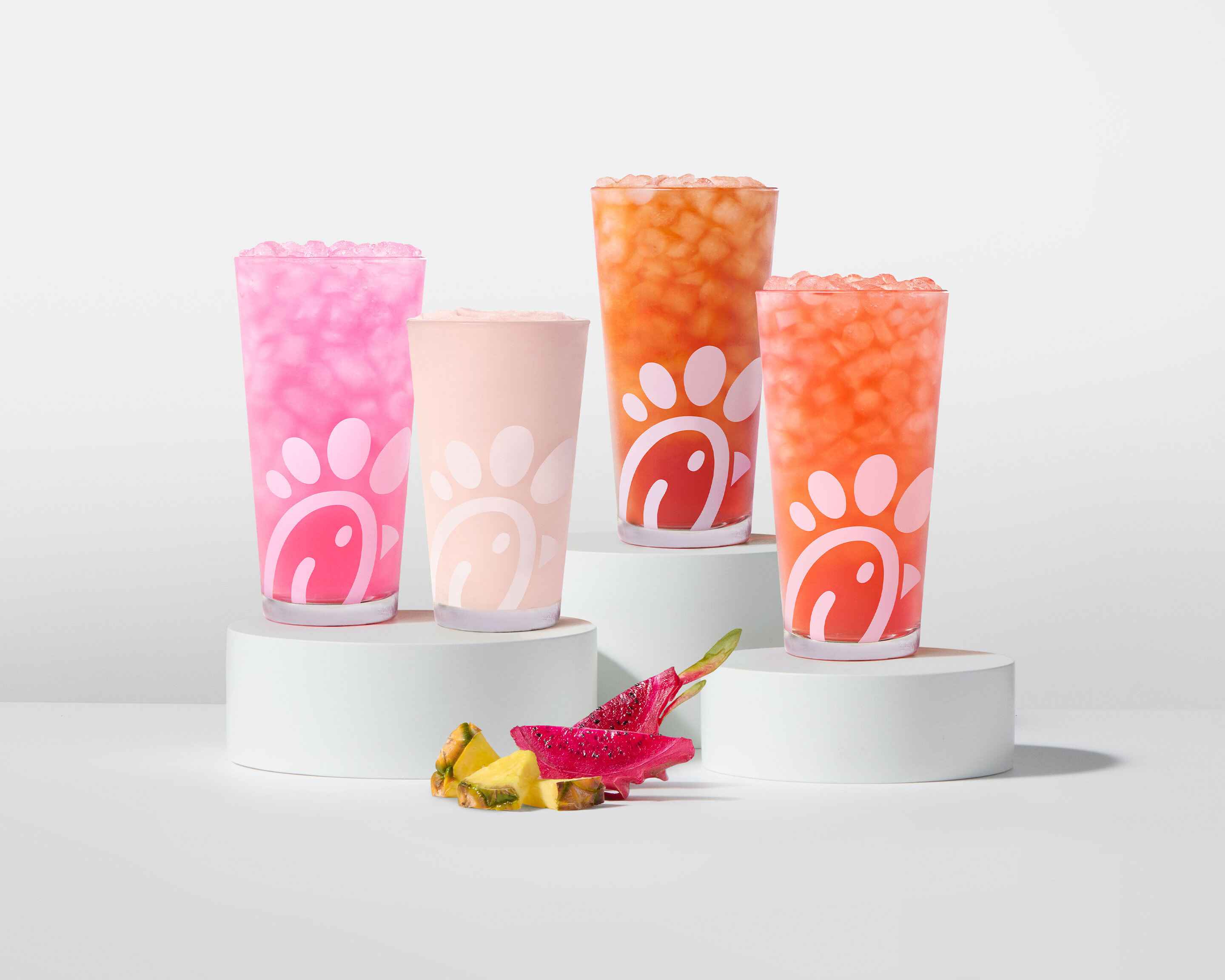 A glass of a Pineapple Dragonfruit Lemonade, Frosted Lemonade, Iced Tea and Sunjoy® on white stands with pieces of pineapple and dragonfruit next to them on a gray background