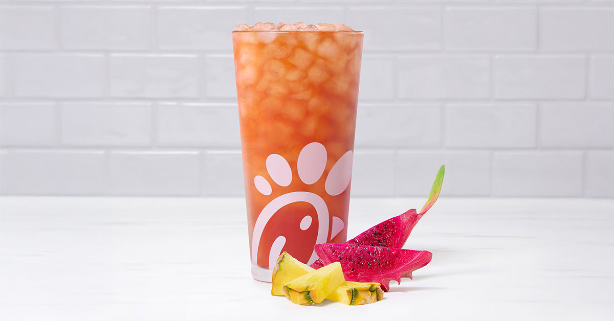 A glass of a Pineapple Dragonfruit Iced Tea with pieces of pineapple and dragonfruit next to it on a tiled background
