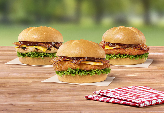 Smokehouse BBQ Bacon Sandwich with Grilled filet, Smokehouse BBQ Bacon Sandwich with Original filet, Smokehouse BBQ Bacon Sandwich with Spicy filet sitting on a table next to a red and white napkin with grass and trees in the background