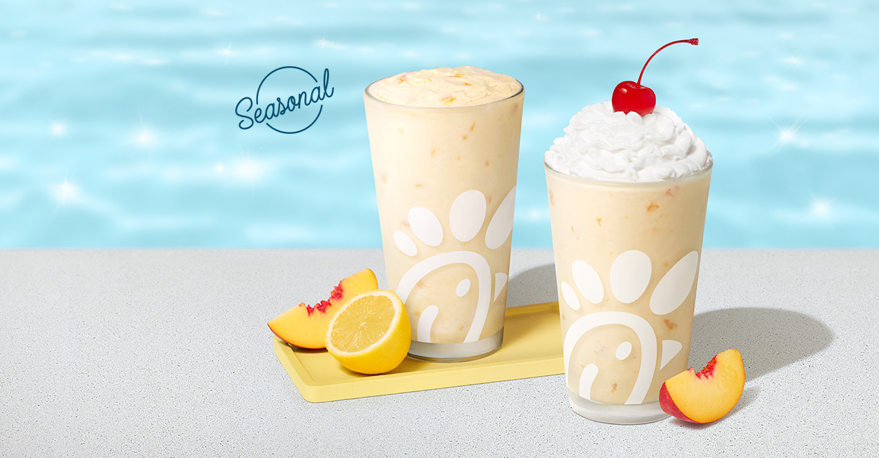Peach Frosted Lemonade in a glass with a peach slice and a lemon half next to it, sitting next to a Peach Milkshake in a glass with a peach slice next to it with a pool in the background