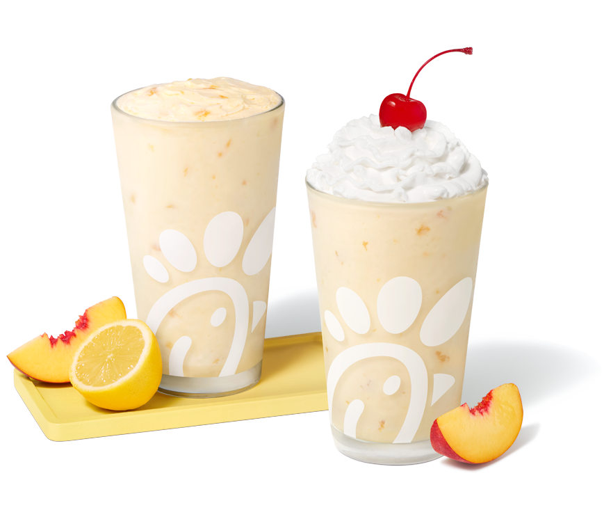 Peach Frosted Lemonade in a glass with a peach slice and a lemon half next to it, sitting next to a Peach Milkshake in a glass with a peach slice next to it